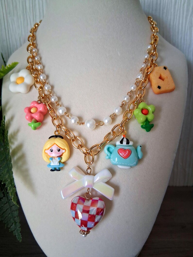 Pu&ograve; includere: Una collana color oro con diversi ciondoli, tra cui un personaggio dei cartoni animati, fiori, una teiera e un biscotto. La collana ha una fila di perle e un ciondolo a forma di cuore con un motivo a scacchi rosso e bianco. Un fiocco bianco &egrave; attaccato al cuore.