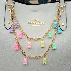 May include: A light-colored handbag with gold-colored hardware and a gold chain strap adorned with colorful gummy bear charms. The charms are in various colors, including pink, purple, blue, and yellow.