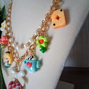 Pu&ograve; includere: Una collana a catena color oro con diversi ciondoli, tra cui una piccola figura di Alice nel Paese delle Meraviglie, una teiera blu, una carta da gioco e un cuore rosso e bianco. La collana presenta anche perle finte bianche e ciondoli a forma di fiore.