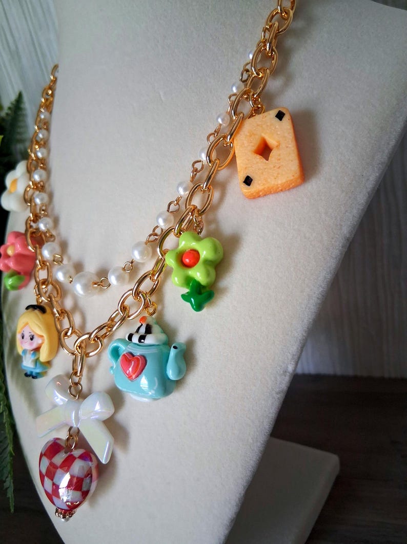 Pu&ograve; includere: Una collana a catena color oro con diversi ciondoli, tra cui una piccola figura di Alice nel Paese delle Meraviglie, una teiera blu, una carta da gioco e un cuore rosso e bianco. La collana presenta anche perle finte bianche e ciondoli a forma di fiore.
