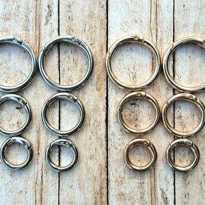 Puede incluir: Un surtido de anillos de resorte de metal plateado y dorado en varios tamaños, dispuestos sobre una superficie de madera clara. Los anillos son circulares con una abertura con bisagras.