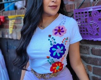 May include: A woman with long black hair is wearing a white t-shirt with colorful embroidered flowers. She is also wearing a purple skirt and a gold belt.