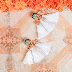 May include: A pair of white tassel earrings with gold accents and colorful mosaic-style round charms. The earrings are on a tiled surface.