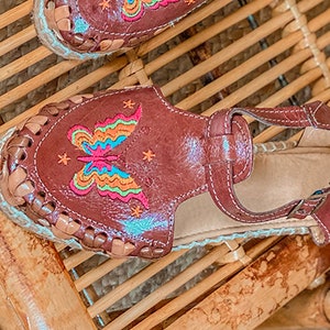 May include: Brown leather sandals with a butterfly embroidered on the front. The butterfly is multi-colored with pink, blue, green, and yellow accents. The sandals have a woven jute sole.