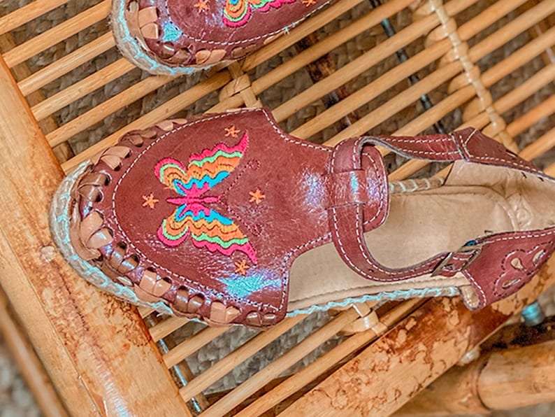 May include: Brown leather sandals with a butterfly embroidered on the front. The butterfly is multi-colored with pink, blue, green, and yellow accents. The sandals have a woven jute sole.