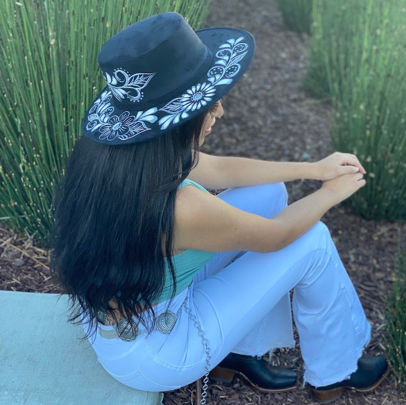 May include: A black felt cowboy hat with a white floral design. The hat is being worn by a person with long black hair and white jeans.