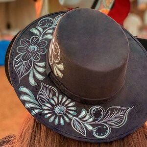 May include: A black felt fedora hat with a wide brim, decorated with a silver floral design. The hat has a flat top and a narrow band around the crown.