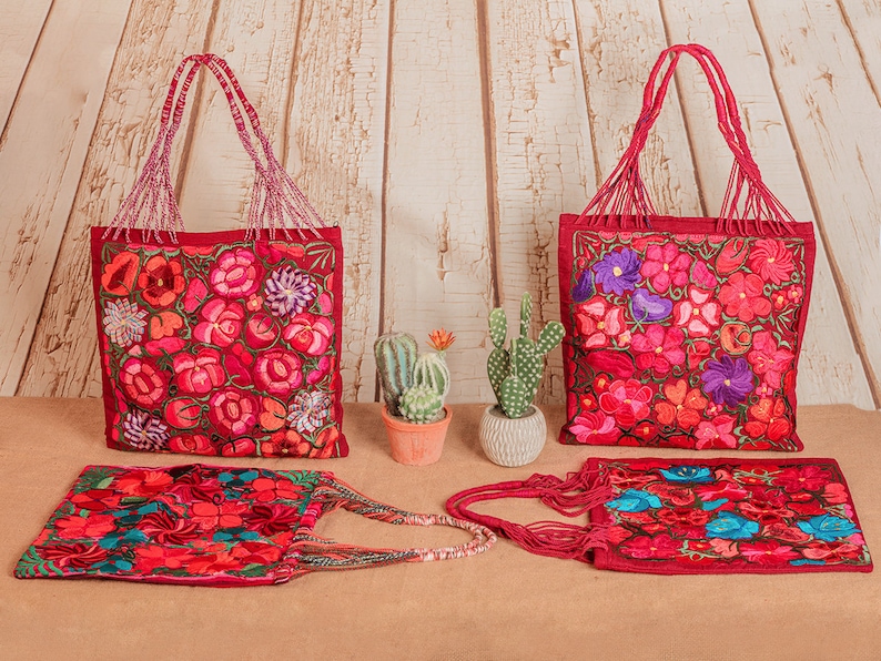 May include: Four red tote bags with colorful floral embroidery. The bags have long, beaded handles. Two of the bags are standing upright, and two are lying flat. The bags are on a light brown surface in front of a white wood background.
