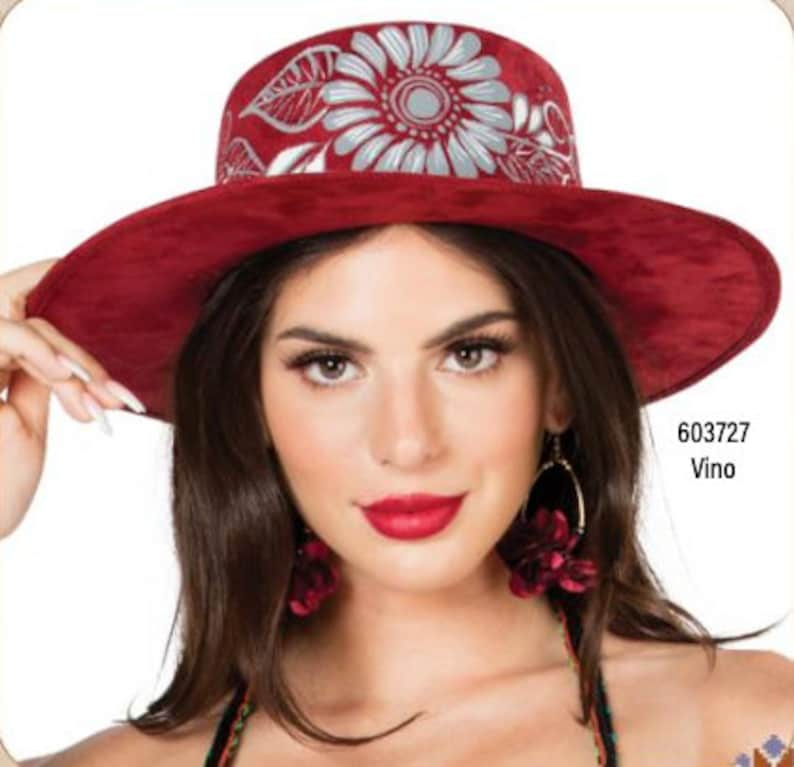 May include: A wide-brimmed hat in red with a white floral design. The hat has a flat top and a wide brim. The hat is made from a soft, suede-like material. The hat is being worn by a person with long brown hair and red lipstick.