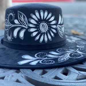 May include: A black wide-brimmed hat with a white floral design painted on the crown and brim.