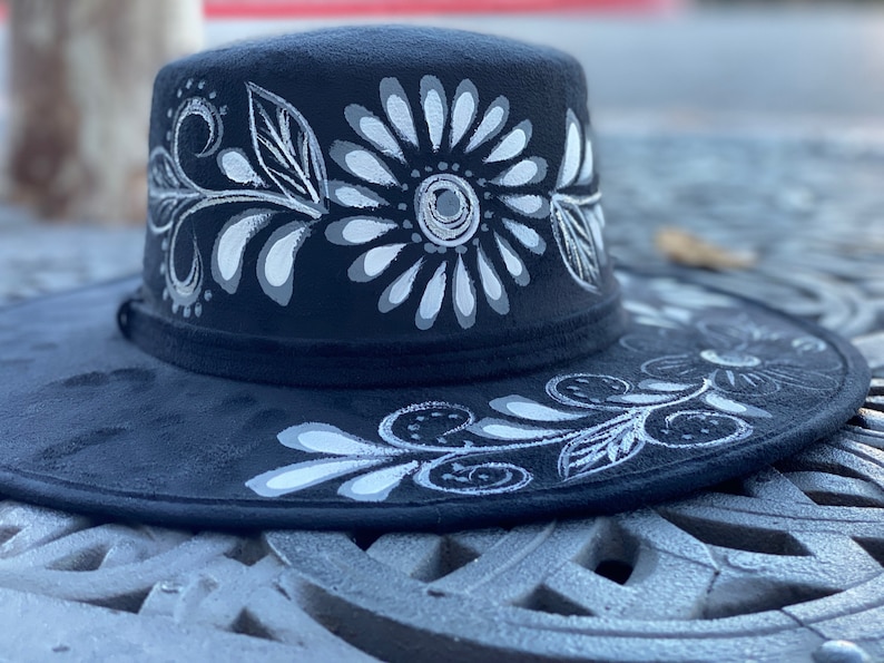 May include: A black wide-brimmed hat with a white floral design painted on the crown and brim.