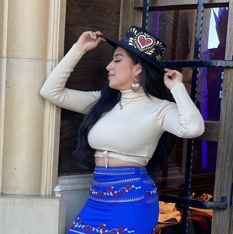 May include: A woman wearing a cream-coloured turtleneck sweater with a black hat featuring a red heart design and gold accents. She is also wearing a blue skirt with red floral embroidery and gold accents.