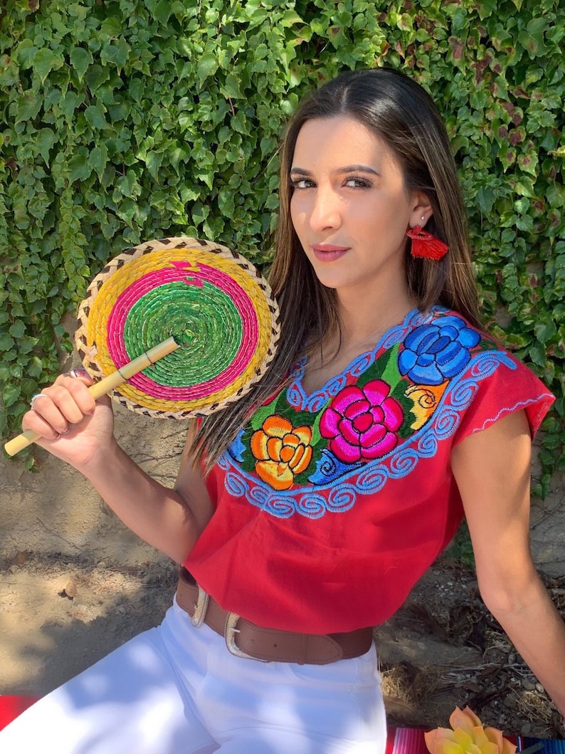May include: A woman wearing a red embroidered blouse with colorful flowers and a brown belt. She is holding a woven fan with a green, yellow, and pink design.