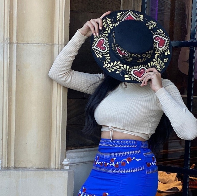 May include: A black hat with gold and red painted hearts and floral designs. The hat is worn by a person with long black hair and a cream coloured top.