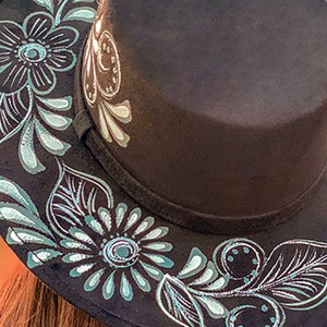 May include: A black fedora hat with a silver and teal floral design painted around the brim.
