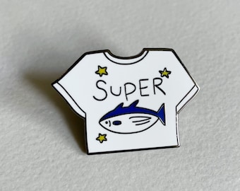 Könnte beinhalten: Weißer Emaille-Pin in Form eines T-Shirts mit dem Wort "SUPER" und einem blauen Fisch darauf. Um das Design sind vier gelbe Sterne angeordnet.