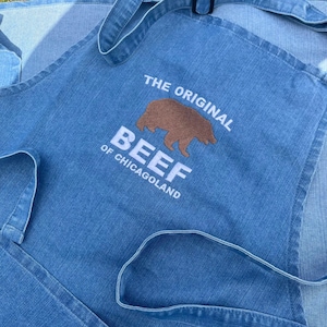 May include: A blue denim apron with the text "The Original Beef of Chicagoland" embroidered in white. A brown bear is embroidered above the text.