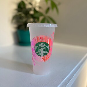 May include: A white reusable cup with a pink and white ombre design. The cup has a green Starbucks logo with the text "Fairy Godmother" in pink and white.