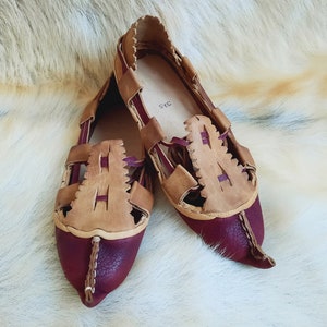 May include: A pair of burgundy leather sandals with tan leather straps and a pointed toe. The sandals have a woven design and are made of leather.