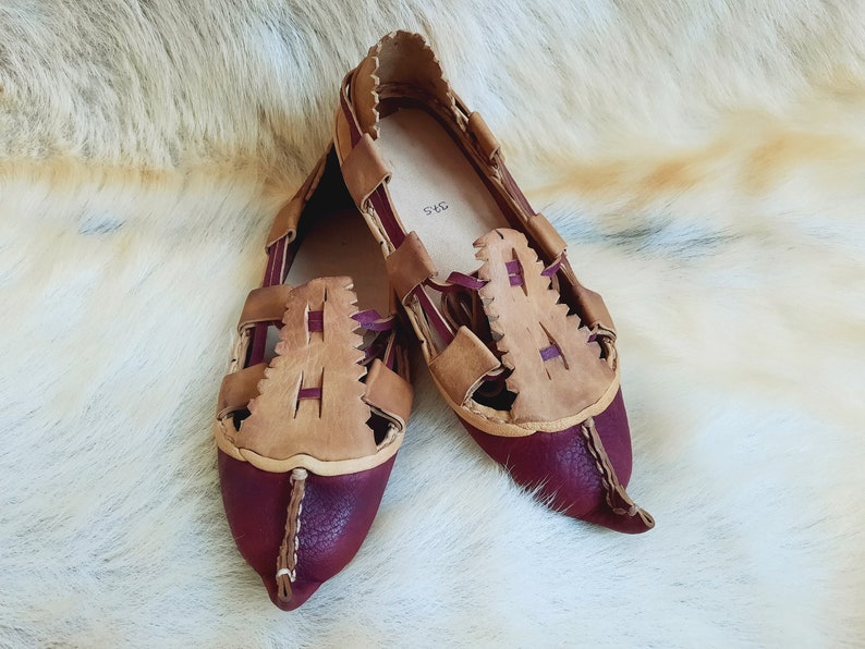 May include: A pair of burgundy leather sandals with tan leather straps and a pointed toe. The sandals have a woven design and are made of leather.