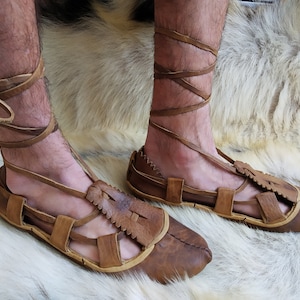 May include: A pair of brown leather sandals with laces that wrap around the ankles. The sandals have a simple design with a few straps and a pointed toe.