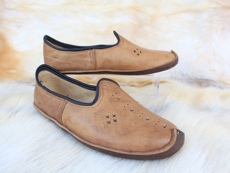 Pu&ograve; includere: Un paio di scarpe marroni in pelle da infilare con bordi neri e ritagli decorativi. Le scarpe hanno una punta arrotondata e una suola piatta.