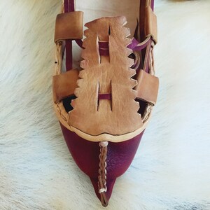 May include: Close-up of a burgundy leather sandal with a brown leather strap and a decorative design. The sandal has a pointed toe and a woven strap.