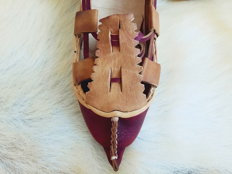 May include: Close-up of a burgundy leather sandal with a brown leather strap and a decorative design. The sandal has a pointed toe and a woven strap.
