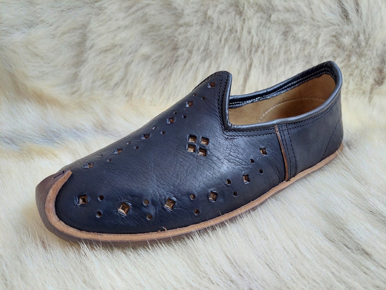 K&ouml;nnte beinhalten: Schwarze Lederschuhe zum Hineinschl&uuml;pfen mit einem dekorativen Ausschnittmuster. Die Schuhe haben eine spitze Zehenpartie und eine braune Ledersohle.
