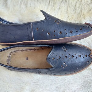 K&ouml;nnte beinhalten: Paar schwarze Slipper aus Leder mit perforiertem Design und braunen Sohlen. Die Schuhe haben eine spitze Zehenpartie und einen schlichten, minimalistischen Stil.