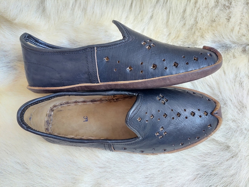 K&ouml;nnte beinhalten: Paar schwarze Slipper aus Leder mit perforiertem Design und braunen Sohlen. Die Schuhe haben eine spitze Zehenpartie und einen schlichten, minimalistischen Stil.