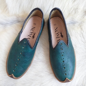 May include: A pair of teal blue leather slip-on shoes with a black trim and a brown sole. The shoes have a perforated design and the brand name "NOMAD" is printed on the inside of each shoe.