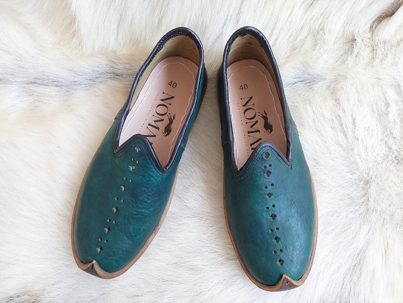 May include: A pair of teal blue leather slip-on shoes with a black trim and a brown sole. The shoes have a perforated design and the brand name "NOMAD" is printed on the inside of each shoe.
