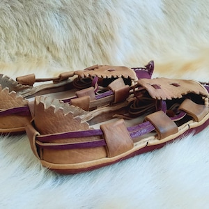 May include: A pair of brown leather sandals with purple straps. The sandals have a unique design with a jagged edge around the opening.