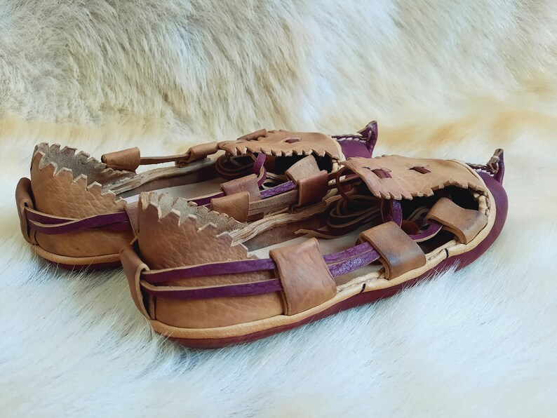 May include: A pair of brown leather sandals with purple straps. The sandals have a unique design with a jagged edge around the opening.