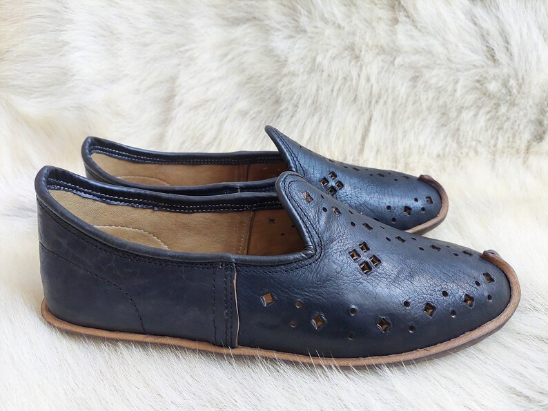 K&ouml;nnte beinhalten: Paar schwarze Leder-Slipper mit perforiertem Design. Die Schuhe haben eine spitze Zehenpartie und eine flache Sohle.
