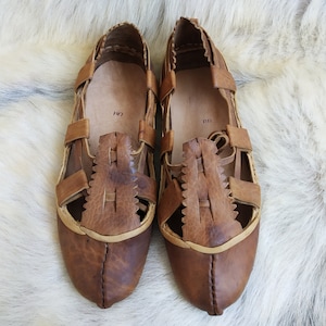 May include: A pair of brown leather sandals with cutouts and a criss-cross strap design. The sandals have a pointed toe and a flat sole.