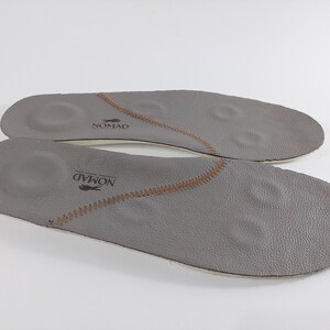 May include: A pair of gray leather insoles with brown stitching and the word "NOMAD" printed on them.
