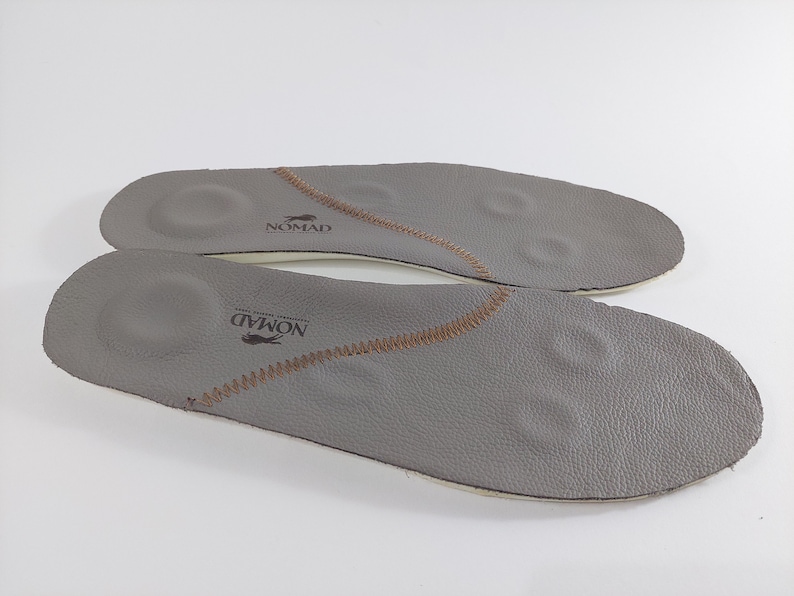 May include: A pair of gray leather insoles with brown stitching and the word "NOMAD" printed on them.