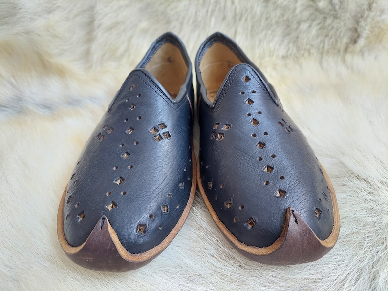K&ouml;nnte beinhalten: Ein Paar schwarze Slipper aus Leder mit einem dekorativen Muster aus kleinen L&ouml;chern und Diamanten. Die Schuhe haben einen braunen Lederbesatz an den R&auml;ndern.