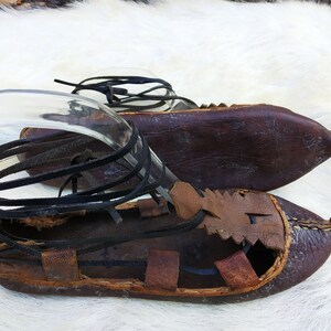 May include: A pair of brown leather sandals with black leather straps. The sandals have a pointed toe and are tied up the ankle.