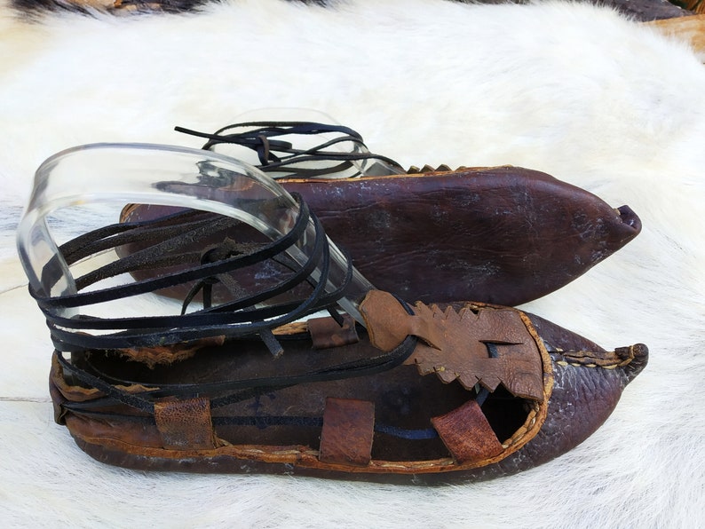 May include: A pair of brown leather sandals with black leather straps. The sandals have a pointed toe and are tied up the ankle.