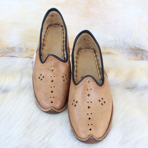 Pu&ograve; includere: Un paio di pantofole in pelle marrone con bordi neri e ritagli decorativi. Le pantofole sono su uno sfondo di pelliccia bianca.