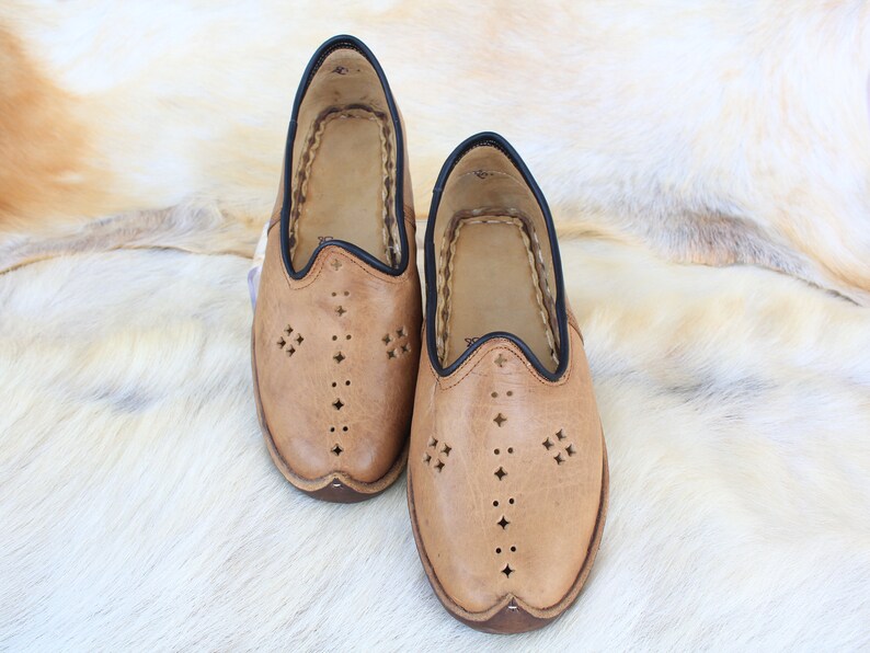 Pu&ograve; includere: Un paio di pantofole in pelle marrone con bordi neri e ritagli decorativi. Le pantofole sono su uno sfondo di pelliccia bianca.