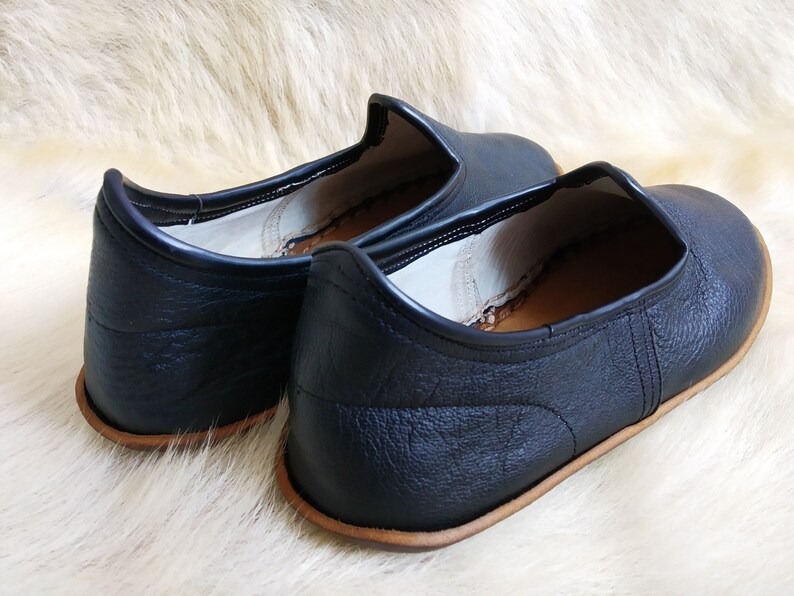 May include: Pair of black leather slip-on shoes with a brown leather sole. The shoes have a simple design and are made of a soft, supple leather.