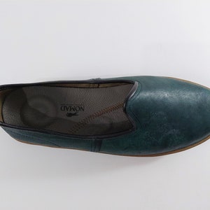 May include: A pair of teal blue leather slip-on shoes with a pointed toe and a brown leather sole. The shoes have a leather insole with the word "NOMAD" printed on it.