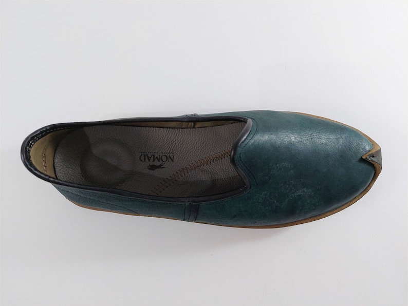 May include: A pair of teal blue leather slip-on shoes with a pointed toe and a brown leather sole. The shoes have a leather insole with the word "NOMAD" printed on it.