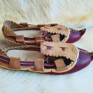 May include: A pair of brown leather sandals with burgundy accents. The sandals have a unique design with multiple straps and a pointed toe.