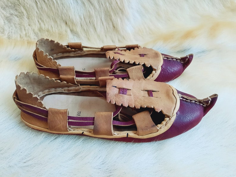 May include: A pair of brown leather sandals with burgundy accents. The sandals have a unique design with multiple straps and a pointed toe.