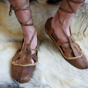May include: A pair of brown leather sandals with laces that wrap around the ankles. The sandals have a simple design with cutouts and are worn on a white furry surface.